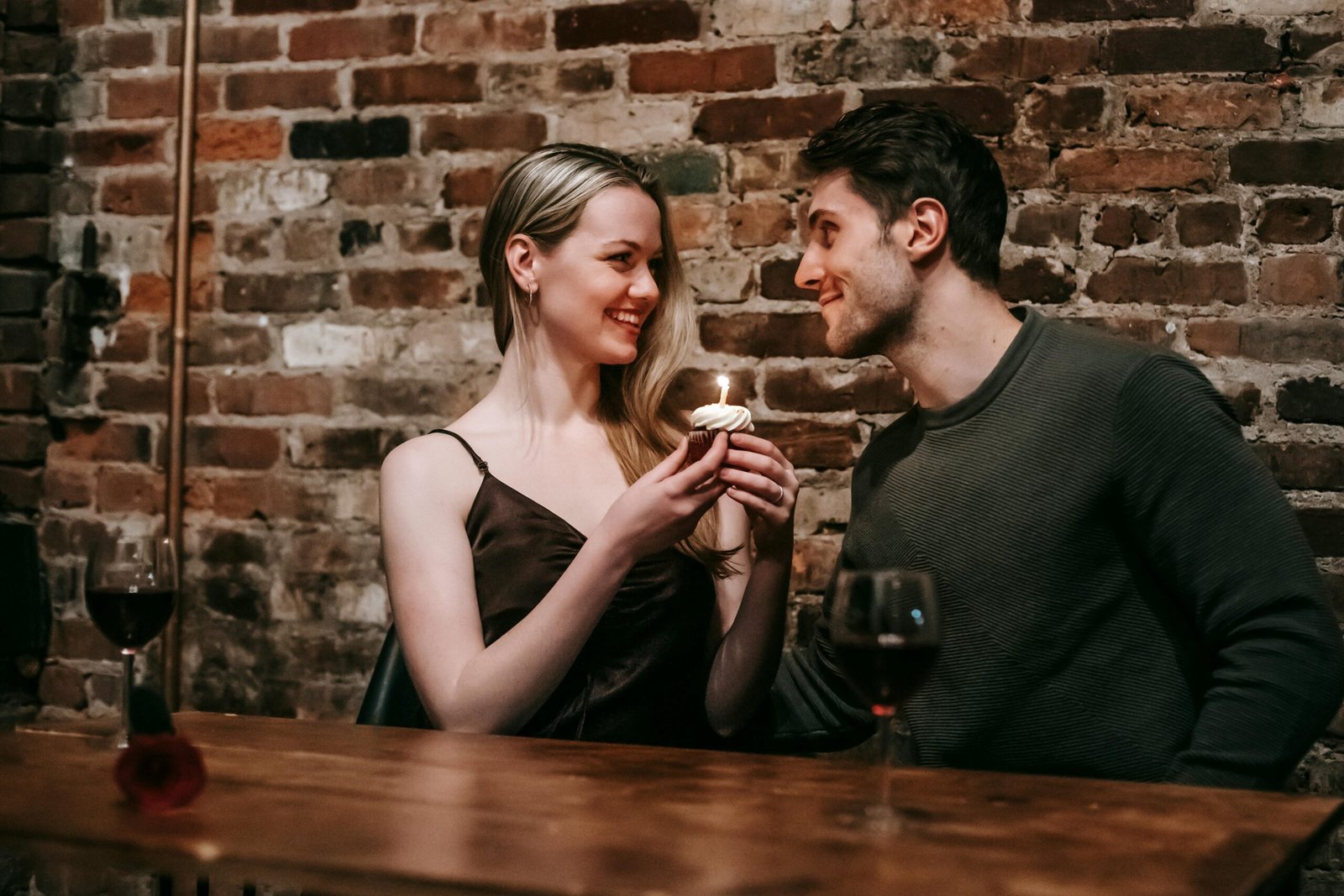 Side view of loving couple sitting near brick wall at wooden table with cupcake with burning candle looking at each other while celebrating anniversary in cafe