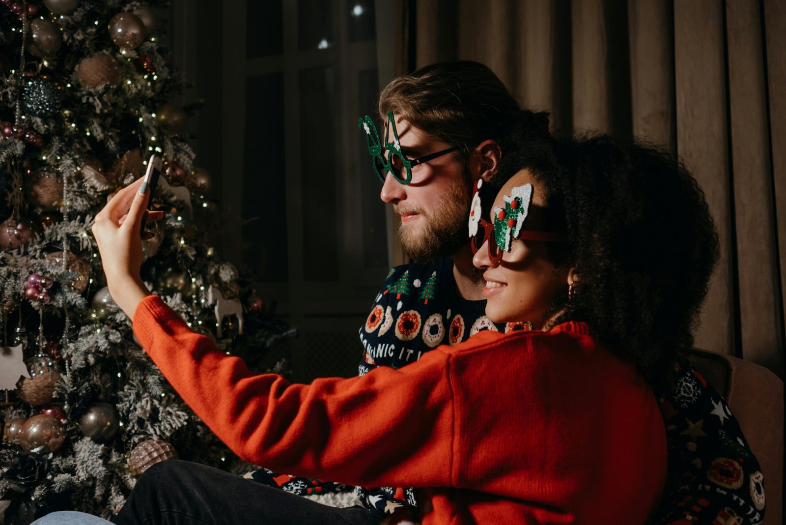 A couple wearing festive glasses enjoys a cozy indoor selfie by a decorated Christmas tree at night.